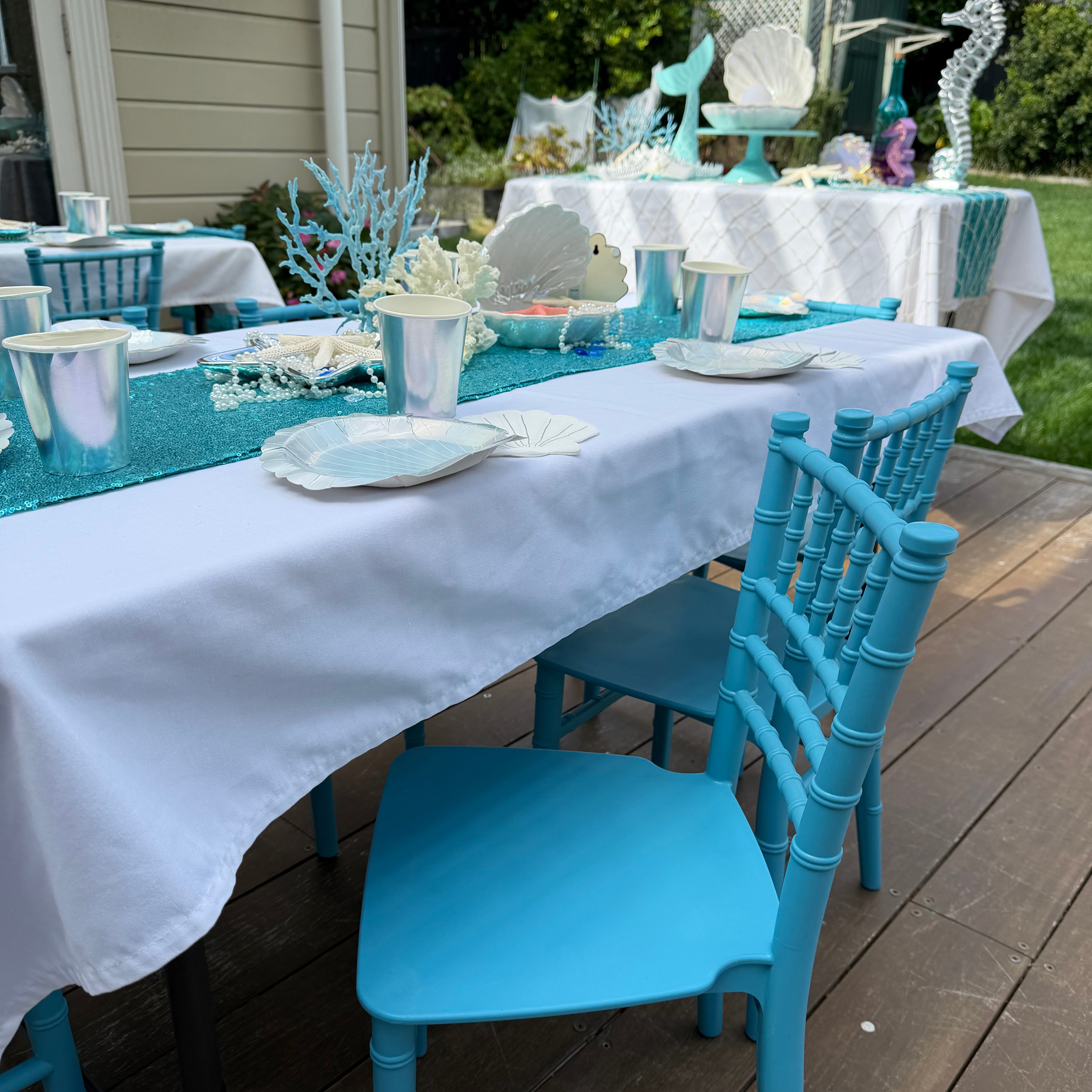 Outdoor setting with blue chairs and white tablecloths on a wooden deck.