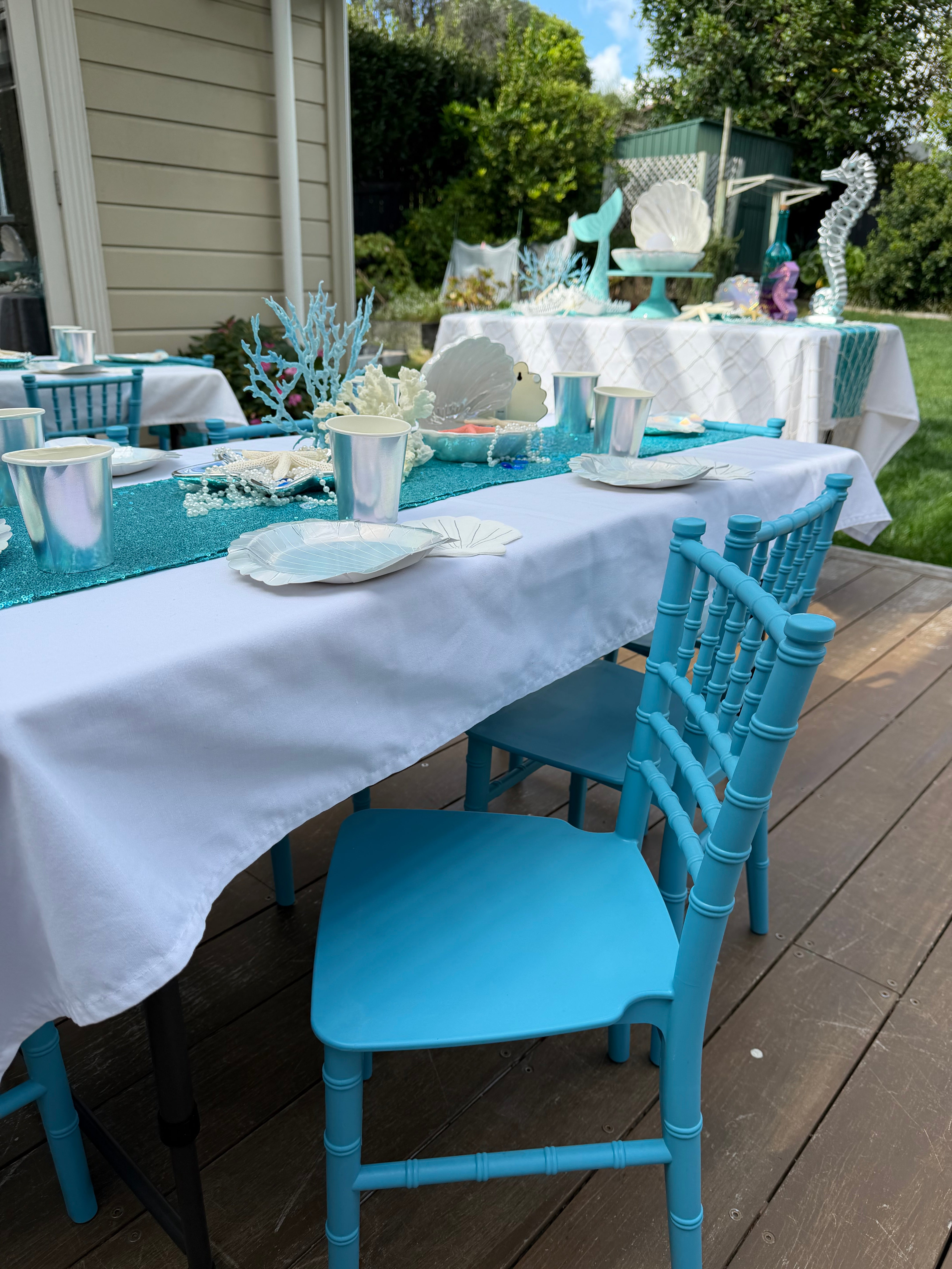 Outdoor setting with blue chairs and white tablecloths on a wooden deck.