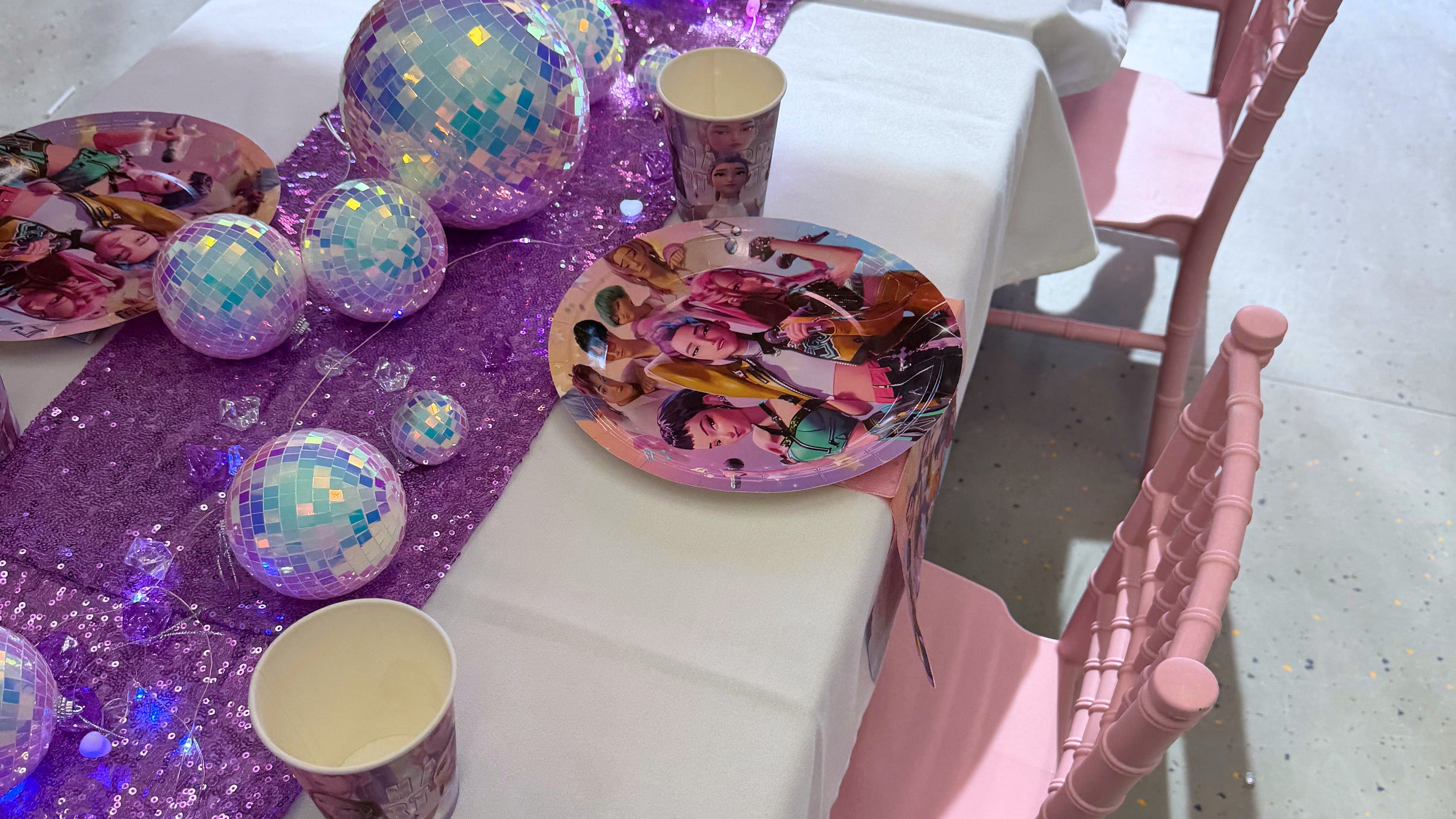 Children's birthday party table setup with pink chairs, colorful plates, and a purple tablecloth.