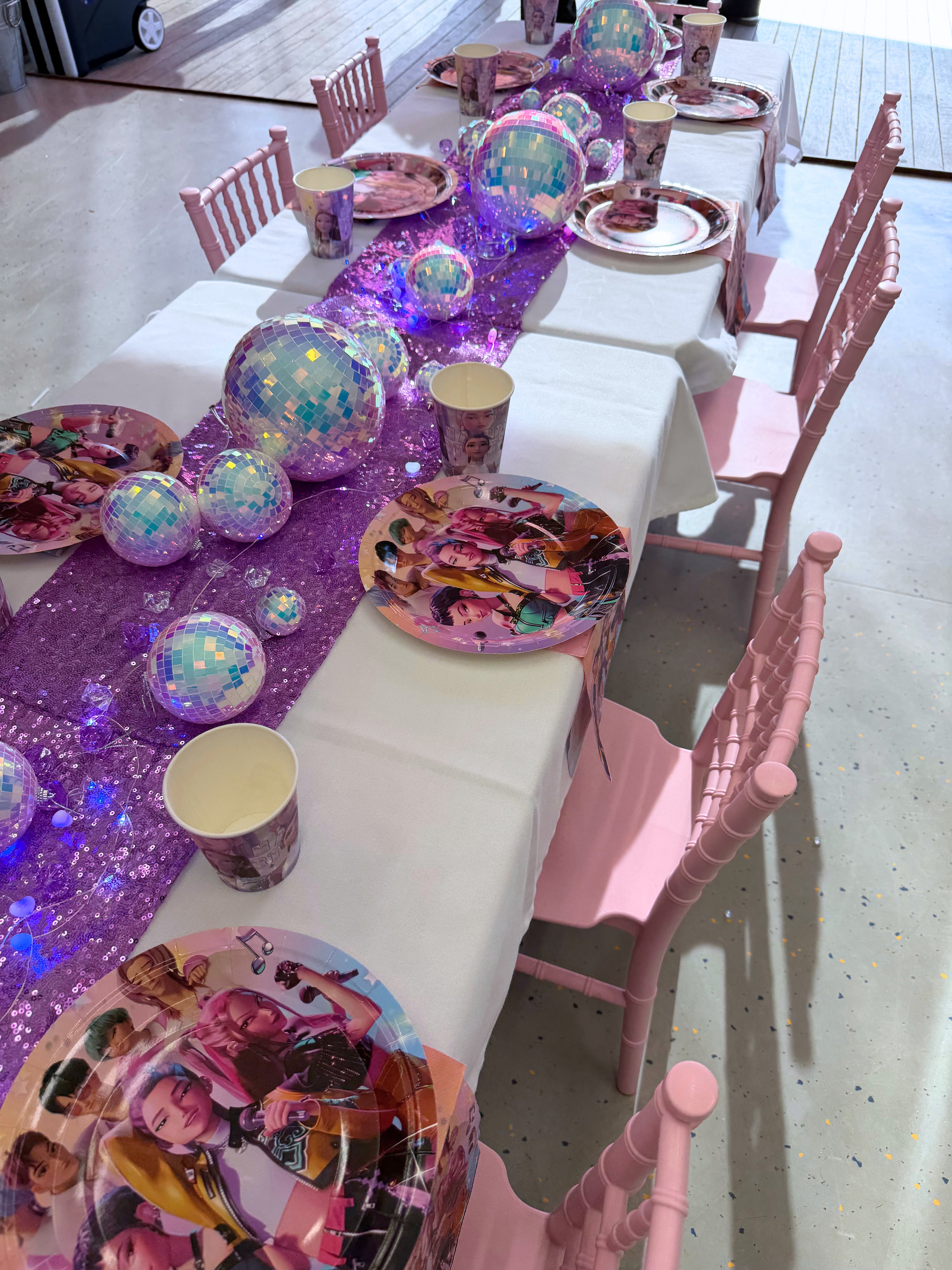 Children's birthday party table setup with pink chairs, colorful plates, and a purple tablecloth.