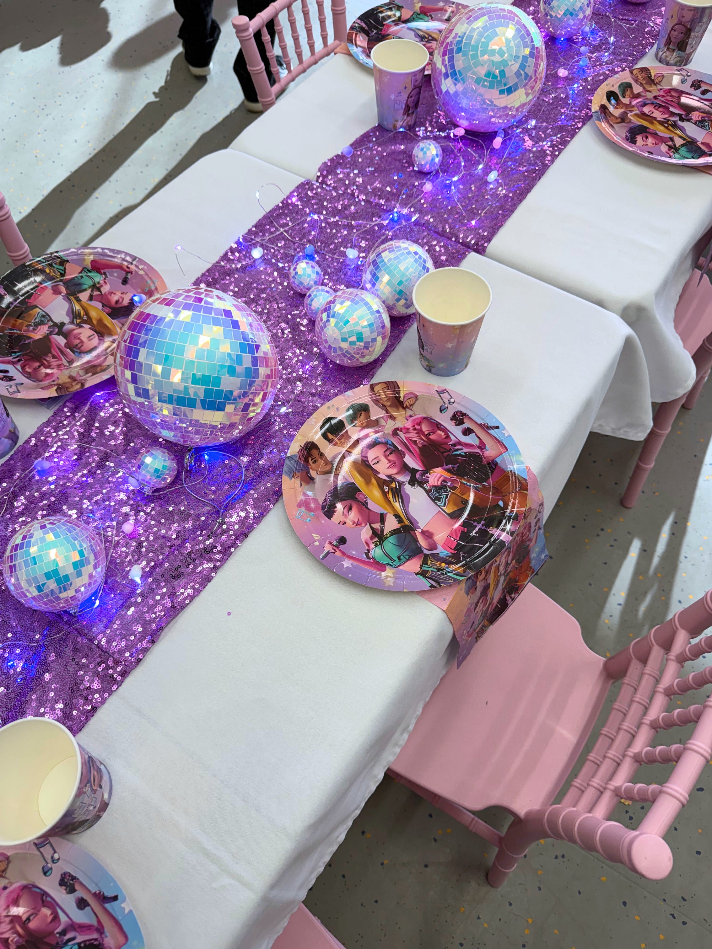 Children's party table setup with purple glitter tablecloth, disco balls, and themed plates.