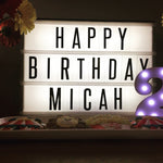 Lightbox with 'Happy Birthday Micah' text, surrounded by toys and flowers on a dark background