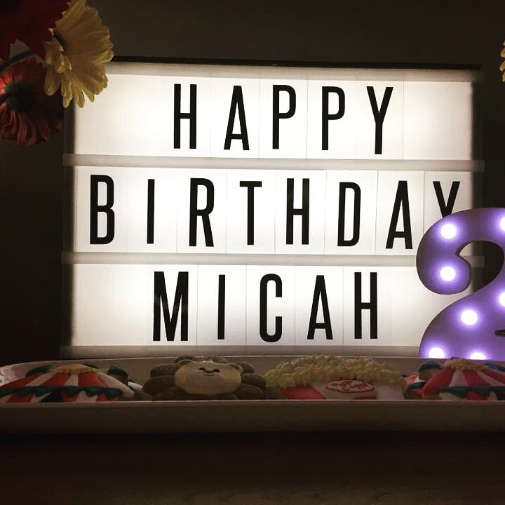 Lightbox with 'Happy Birthday Micah' text, surrounded by toys and flowers on a dark background