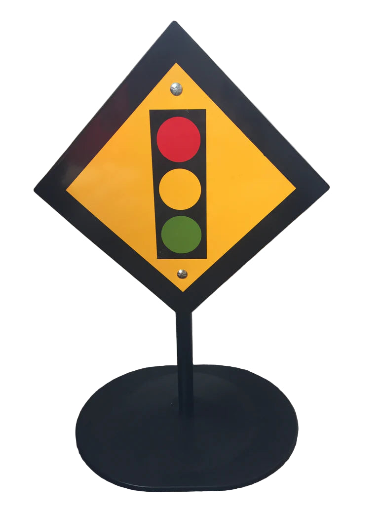 Diamond-shaped traffic sign with red, yellow, and green lights on a black stand against a white wall.