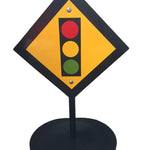 Diamond-shaped traffic sign with red, yellow, and green lights on a black stand against a white wall.